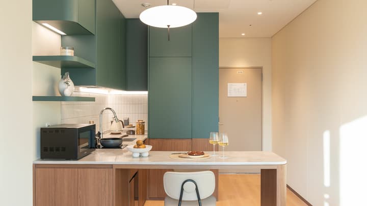 Close-up of the stylish kitchenette with bold dark green cabinets and floating shelves, integrated microwave, and a counter that extends into a two-seater dining bar in the one-bedroom suite.