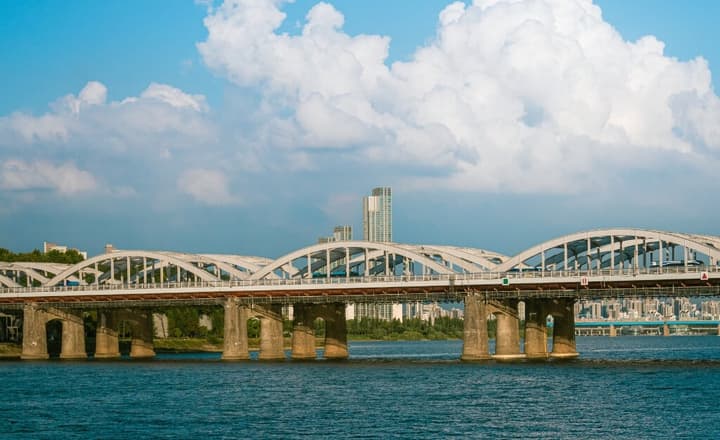 Wide shot of a large, arched steel bridge crossing the Han River in Seoul, supported by thick concrete pillars, with the modern city skyline visible on the far bank under a cloudy sky.