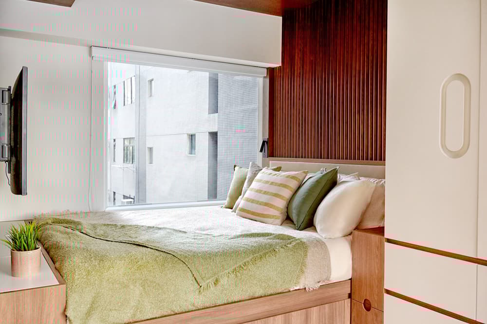 Cozy, modern bedroom featuring a light wood platform bed with green and striped pillows, a warm wooden slatted accent wall, and a large horizontal window.