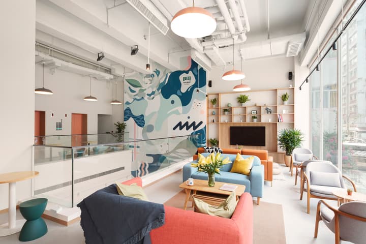 A bright, two-story co-living communal lounge with contemporary orange and blue couches, a large abstract wall mural, and natural light from tall windows.