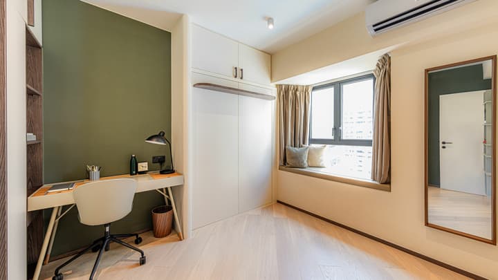 Flexible room set up as a study with a white desk against a deep green accent wall, built-in storage, and a cushioned window seat.