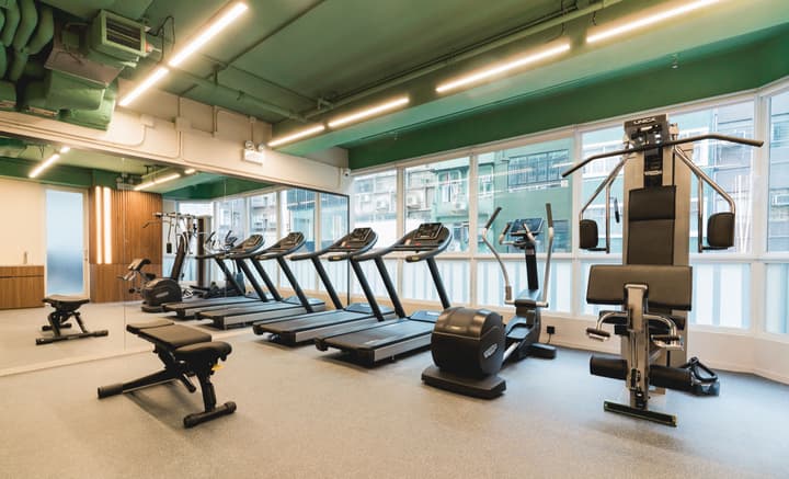 Well-equipped fitness center with a bright green ceiling, featuring a row of treadmills, an elliptical machine, a multi-station weight machine, and free-weight benches, with large mirrors and windows overlooking a city.