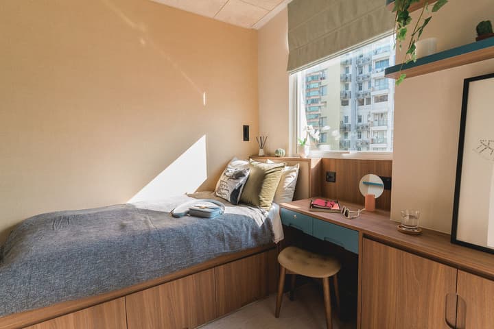 A compact, modern bedroom setup in an Urban Suite at Weave Suites – Central West, featuring a single bed with under-bed storage, a built-in desk with a light blue drawer unit, and a window.