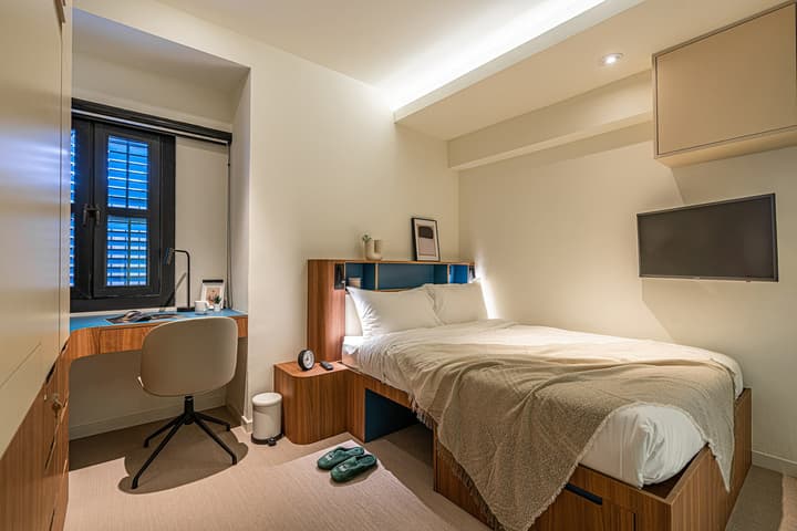 Compact premium suite bedroom featuring a queen bed with integrated under-bed storage, a built-in desk by the window, and a wall-mounted television.