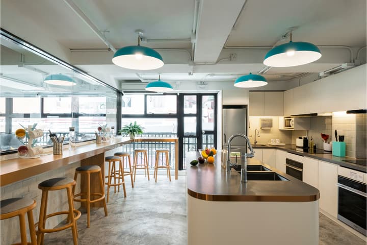 Bright, modern communal kitchen featuring white cabinetry, a large central island with a sink, and a high counter with wooden bar stools leading to a doorway for an outdoor area, all illuminated by turquoise pendant lamps.