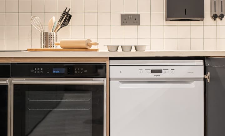 Close-up of a modern kitchen countertop and backsplash of white square tiles, showing a black built-in oven next to a white integrated dishwasher. Kitchen utensils and a wooden rolling pin sit on the counter above.