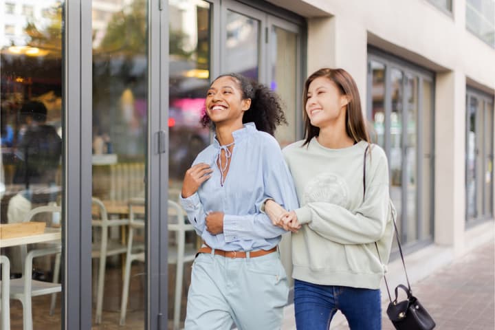 Two young women, one with curly hair and one with straight hair, smiling brightly as they walk arm-in-arm past a glass-fronted cafe or storefront.