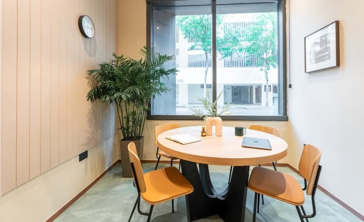 Small meeting room with a round wooden table, four chairs, a large window providing natural light and a view of the street, and a potted plant in the corner.