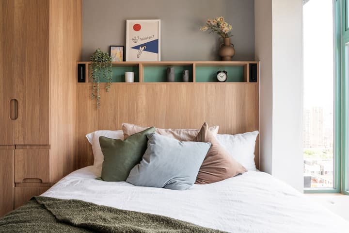 Close-up of a queen-sized bed with white bedding and a mix of colorful pillows. The bed features a built-in wood headboard with an integrated, light-green accented shelf holding artwork, plants, and a clock. Tall wood-grain cabinetry is visible on the left.