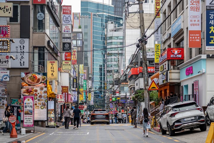 A narrow, busy street in Gangnam, Seoul, lined with buildings covered in a vibrant array of vertical Korean restaurant and shop signs, with pedestrians and cars in the distance.