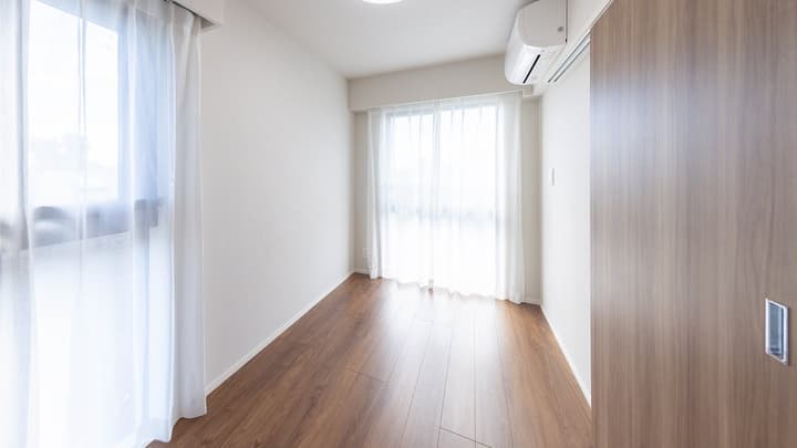 Empty bedroom with light hardwood floors, large windows with sheer curtains providing access to a balcony, a second window, and a wall-mounted air conditioning unit.
