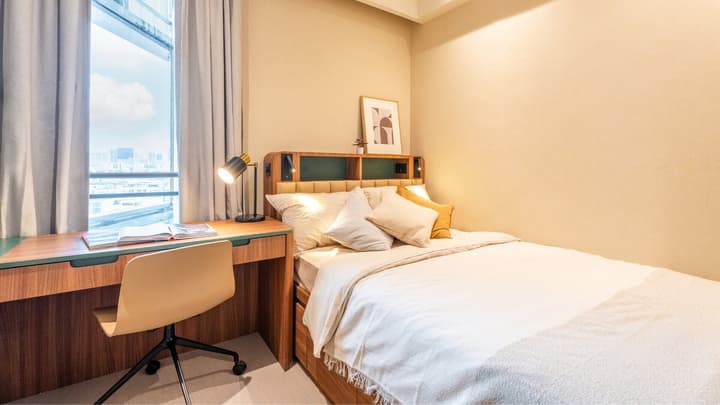 Secondary bedroom with a queen bed, integrated headboard shelving, and a large desk positioned by a window with a city view.
