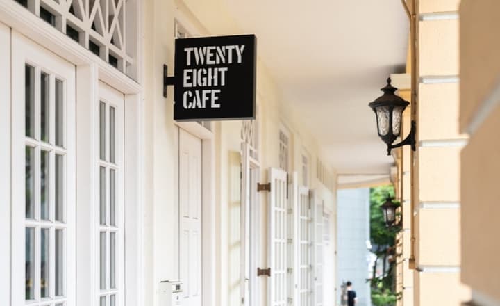 "Twenty Eight Cafe" sign hanging on a classical white building exterior with multiple white French doors along a sheltered walkway.