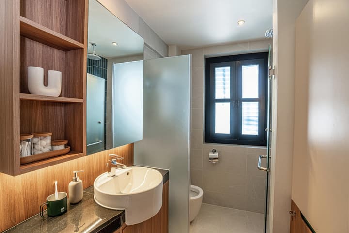 Modern one-bedroom suite bathroom featuring a round vessel sink on a dark counter, wood-look shelving, and a shower area with a window and gray tiled walls.