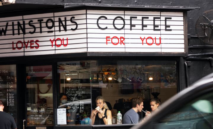 Street view of Winstons Coffee storefront with a marquee-style sign that reads "WINSTONS COFFEE LOVES YOU FOR YOU" in black and red letters, showing people seated inside the window enjoying coffee and food.