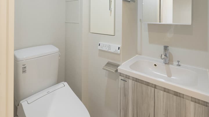 Modern apartment bathroom showing a white washlet-style toilet and a small vanity with a sink, faucet, and wood-grain cabinet underneath.