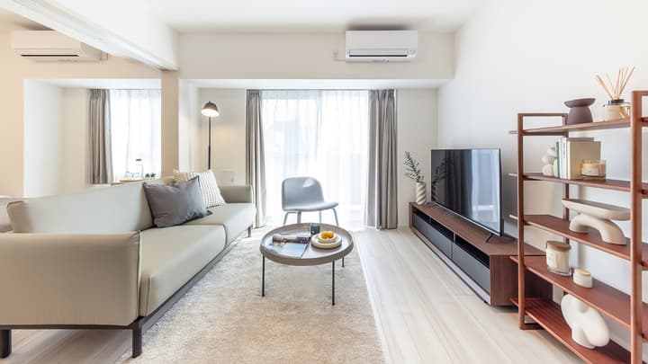 Long, bright living room space featuring a light gray sofa, modern wood shelving unit, and a black TV console under a large, curtained window.