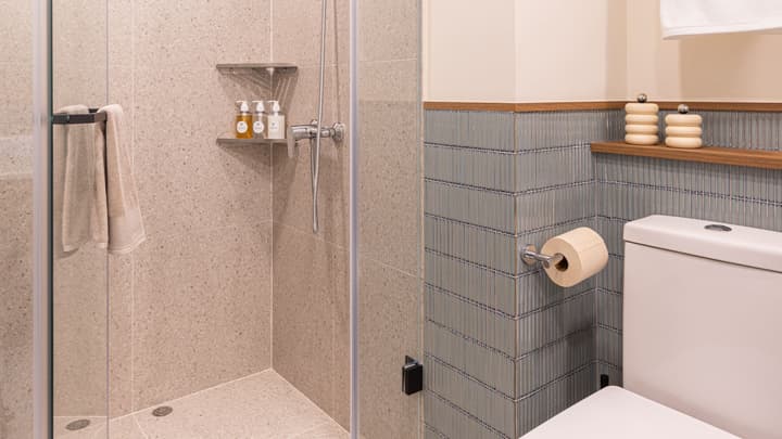 Close-up of a modern bathroom showing the toilet area with vertical blue ridged accent tiles, a wooden shelf, and a glass-enclosed shower with speckled beige terrazzo-style tile.