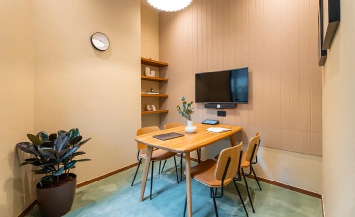 Small meeting room with a light wood table, four chairs, a wall-mounted TV and soundbar for video conferencing, and light acoustic wall panels.