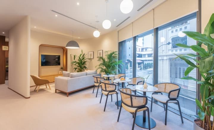 Communal dining and lounge area with a large window, light gray sofa, and several small tables with woven chairs, all well-lit with natural light and hanging globe lights.
