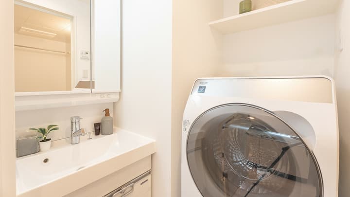 Apartment bathroom vanity area with a sink, mirrored cabinet, and a large, modern front-loading washer-dryer combination unit to the side with a storage shelf above.