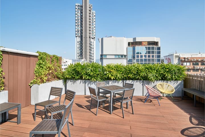 A sunny, landscaped rooftop terrace with wooden decking, a dining table with chairs, and colorful lounge seating, set against a backdrop of modern high-rise buildings.