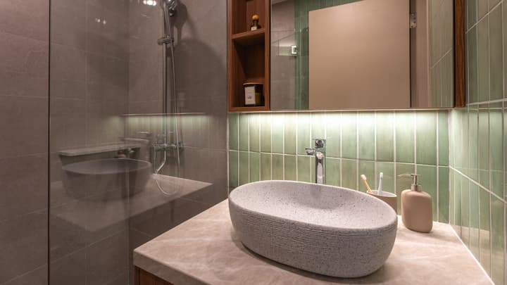 Modern en-suite bathroom with a textured, oval stone basin on a marble vanity, a glass shower partition, and vertical seafoam green subway tiles as a backsplash.