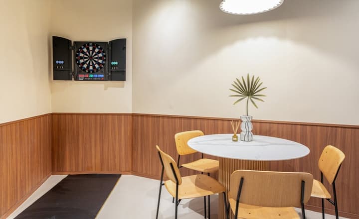 Close-up of an entertainment room corner with a mounted electronic dartboard and a small white marble and wood lounge table with chairs.
