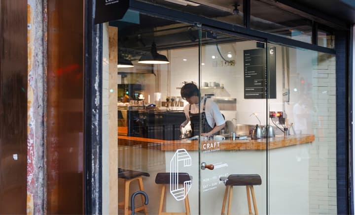View through the glass window of a brightly lit coffee shop, showing a barista working at a wooden counter. The interior features black espresso machines and hanging industrial light fixtures.