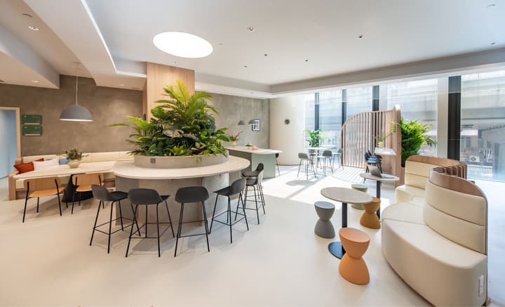 A large, open communal lounge area with multiple seating zones, including cushioned banquette seating, a row of low colorful stools, and a central oval bar counter with black stools, featuring a large planter with tropical plants.