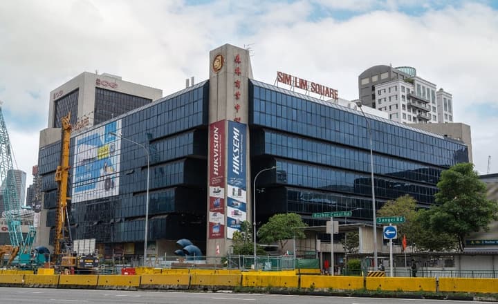 Exterior of Sim Lim Square, a large commercial electronics mall in Singapore, with a dark glass facade and various billboards.