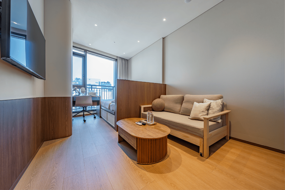 Wide view of a modern studio apartment with a queen bed and a separated sofa bed lounge area featuring a unique adjustable height coffee table, set near a large window.
