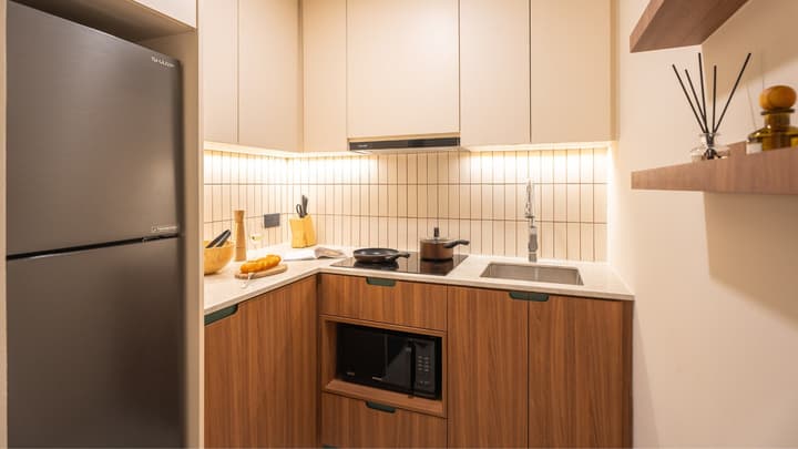 Compact kitchenette with upper and lower beige and walnut cabinets, a black refrigerator, induction cooktop, microwave oven, and beige subway tile backsplash.