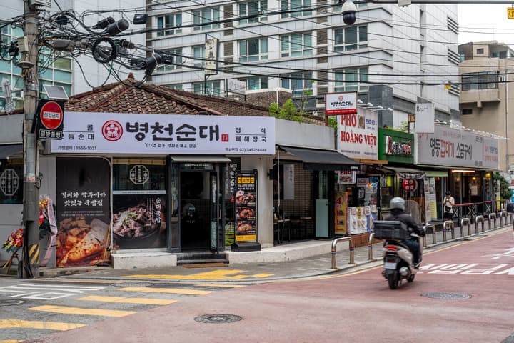 A traditional-style Korean restaurant ("Byeongcheon Sundaeguk") on a street corner, with tiled roofs, white walls, and a menu sign, next to other storefronts like a Pho shop.
