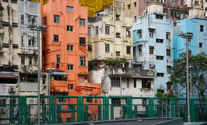 A street-level view of densely packed old apartment buildings in various pastel and earthy colors, including orange and light blue, behind a green chain-link fence.