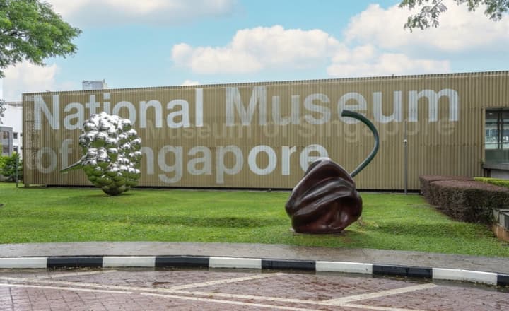 Exterior of the National Museum of Singapore, showing the building signage and large, oversized fruit-themed art sculptures on the front lawn.