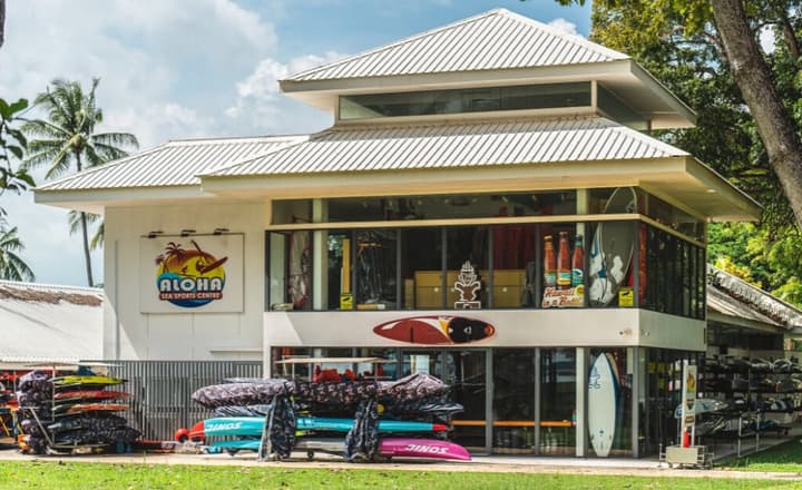 Exterior of the Aloha Sea Sports Centre, a white two-story building stocked with surfboards, kayaks, and other water sports equipment.