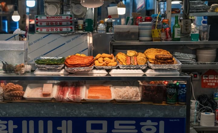 A vibrant Korean street food market stall displaying various pre-cooked items on the top shelf, including fried fish cakes and vegetable pancakes, with raw seafood and canned drinks on the refrigerated bottom shelf.