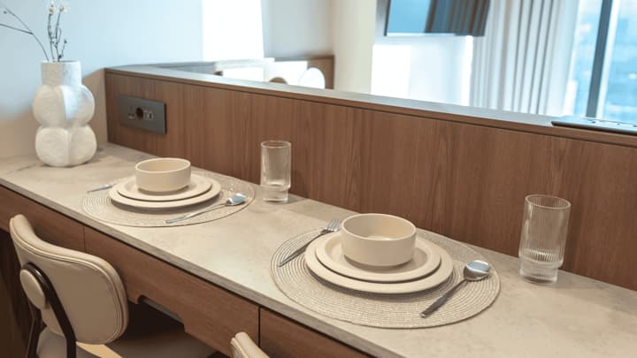 Close-up of a stylish two-person dining counter with contemporary place settings, round placemats, white textured bowls, and clear glasses, next to a decorative white vase.