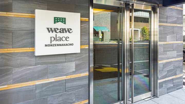 Exterior view of the main entrance to Weave Place Monzennakacho, featuring a contemporary grey slate wall with gold accents, a white sign with the green 'weave place' logo and name, and large glass double doors.