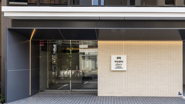 The modern main entrance of Weave Place Asakusa Kaminarimon, featuring a glass door, dark gray paneling, and a light brick wall with the building's logo sign.
