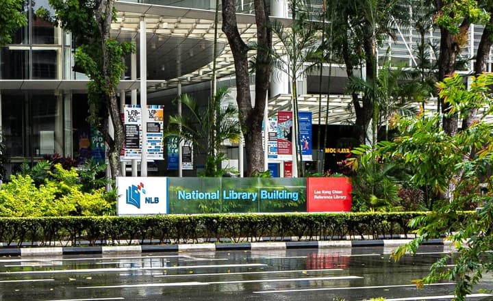 Street view of the National Library Building in Singapore, showing the prominent sign through lush green trees.