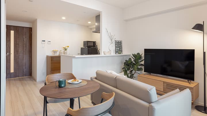 View of the apartment's living space with a light sofa, round dining table set for two, and a bright kitchen counter nook with decor.
