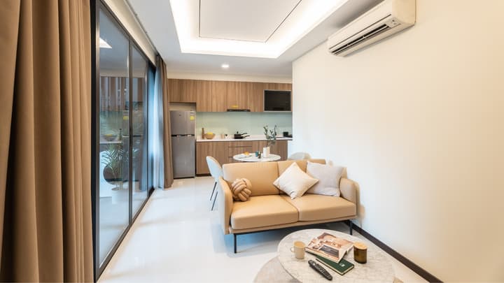 Compact, open-concept living area and kitchen with a light tan sofa, small dining table, and large glass sliding doors to the balcony.