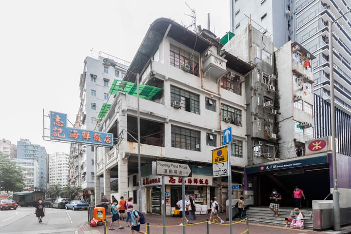A street-level photo in Prince Edward, Hong Kong, showing an old, multi-story building facade next to the purple entrance of the MTR station and people walking along the street.