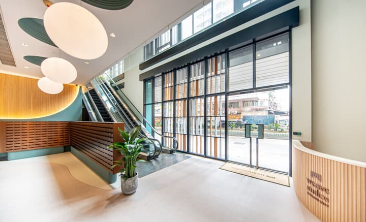 The interior of a modern building lobby with light flooring, a wood-paneled reception desk, and a large escalator leading to the upper floor. The entrance features a tall black-framed glass wall with vertical wooden accents, opening to a city street.