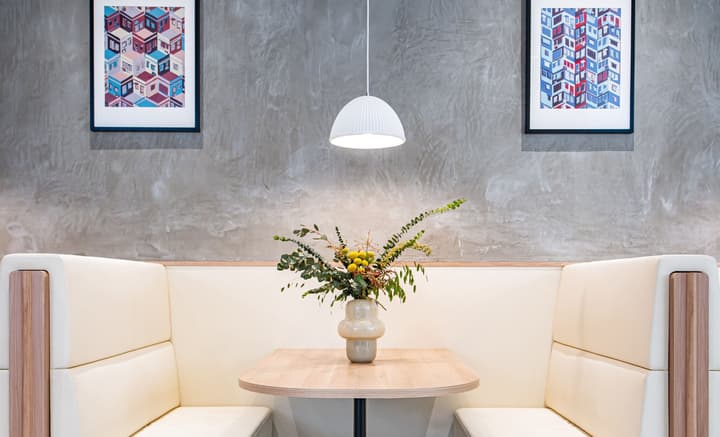 A close-up view of a cozy coworking booth with light beige upholstered seating and a small wooden table. A floral arrangement in a neutral vase sits on the table, framed by a textured gray concrete wall adorned with two modern, colorful geometric artworks.