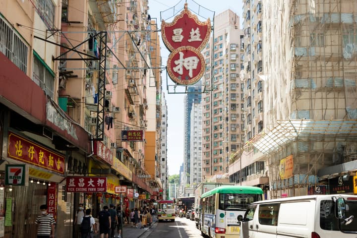 Street-level view of a bustling Hong Kong street with colorful, dense buildings, featuring a distinctive traditional red and gold Chinese pawn shop sign hanging high above a mix of pedestrians and traffic.