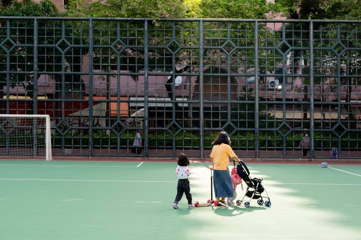 A woman in a yellow shirt pushing a stroller and watching a small child on a scooter on a green, multi-purpose outdoor sports court or playground with a soccer goal.