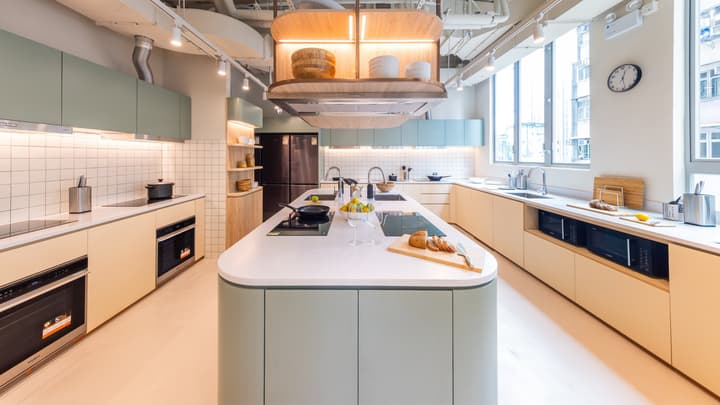 Spacious, modern communal kitchen featuring multiple cooktops and ovens, light green and beige cabinets, a white tile backsplash, and a large central island with sinks.